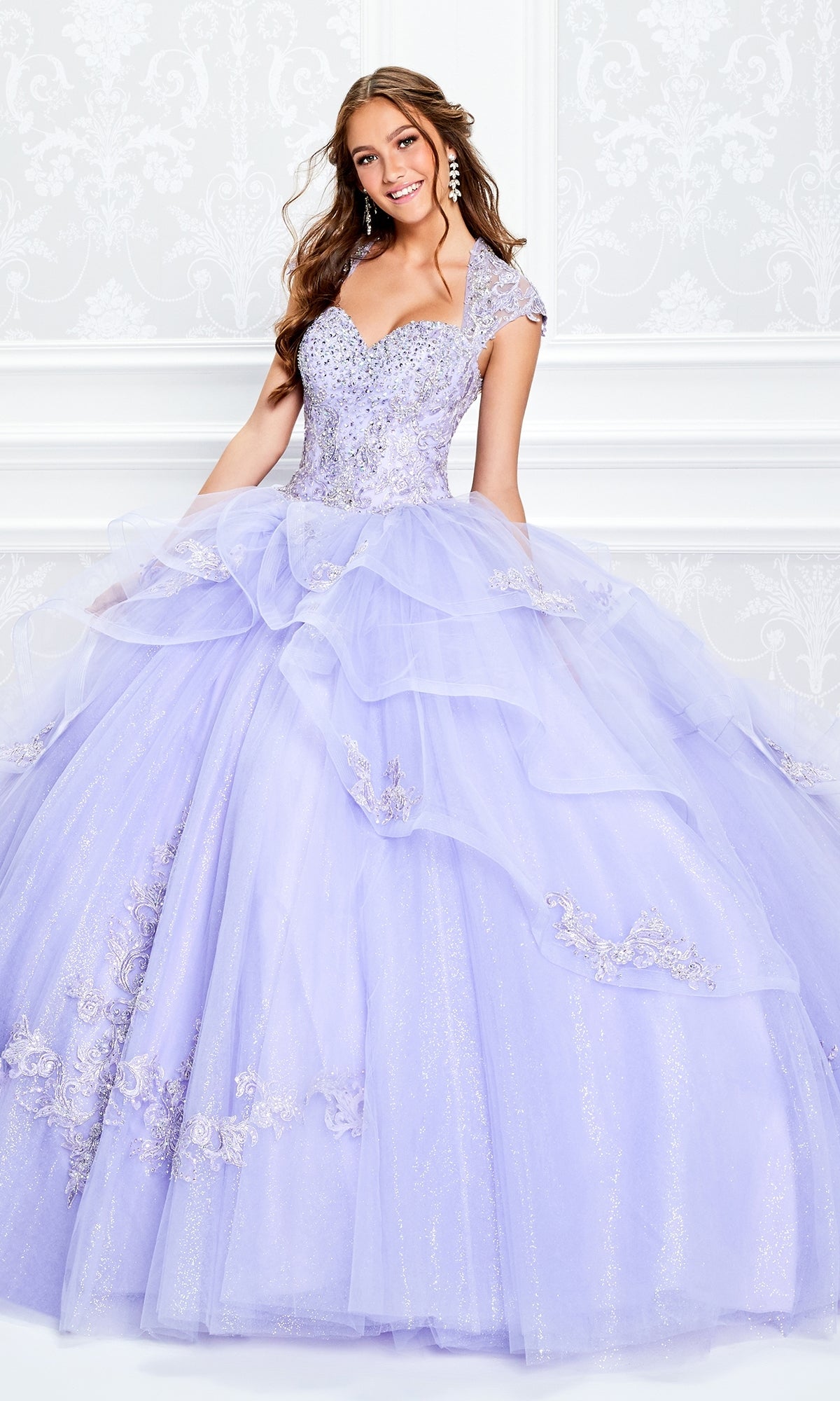 A young woman smiles at the camera in front of a white ornate wall, wearing the Princesa Beaded Lace Quinceañera Dress PR11923, a sparkling lavender ball gown adorned with stunning beaded lace.